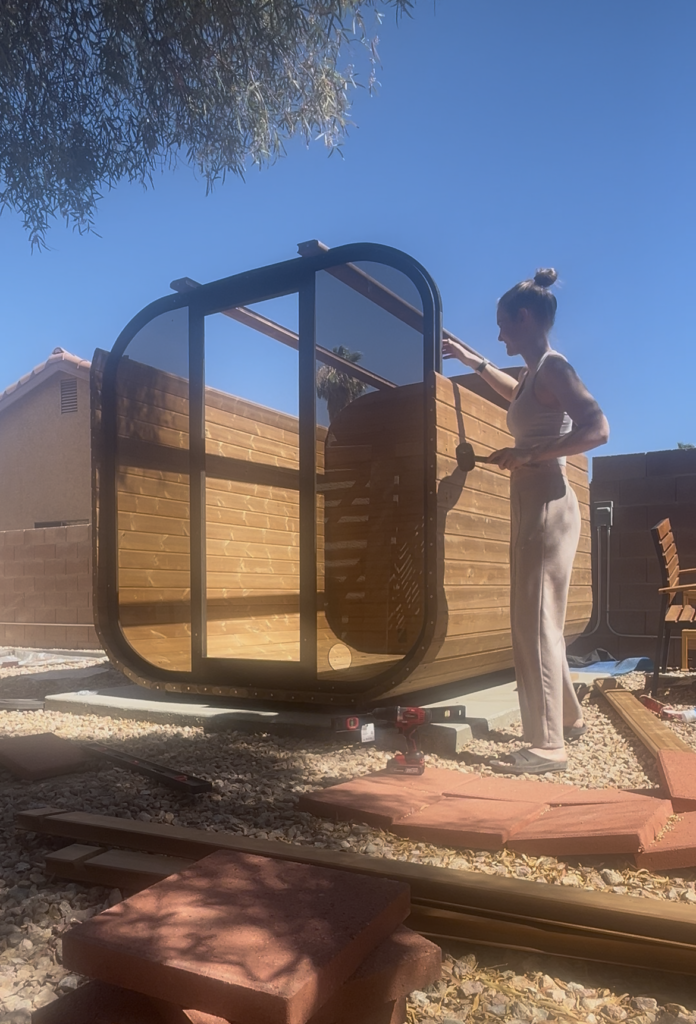Emily standing next to the partially assembled wooden barrel sauna, with its glass front frame and curved wooden walls visible.