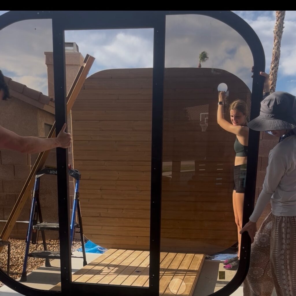 amily members steadying the large glass sauna door frame in front of the partially assembled wooden sauna.