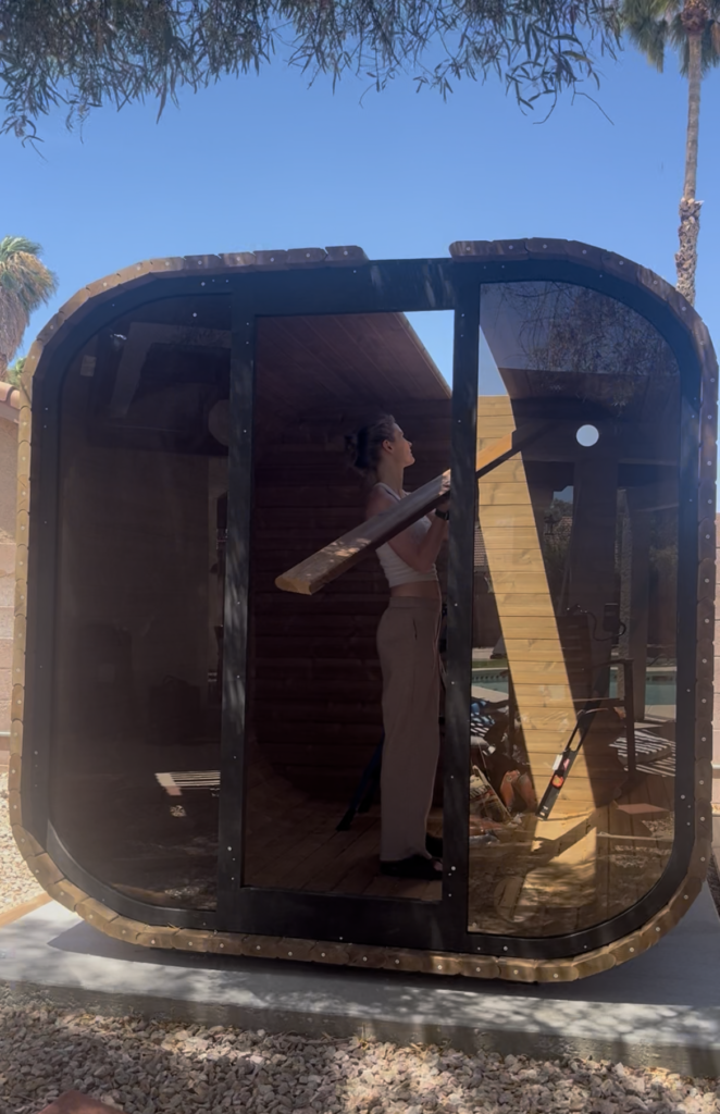 Emily positioning a wooden board inside the unfinished sauna, viewed through the curved front frame.
