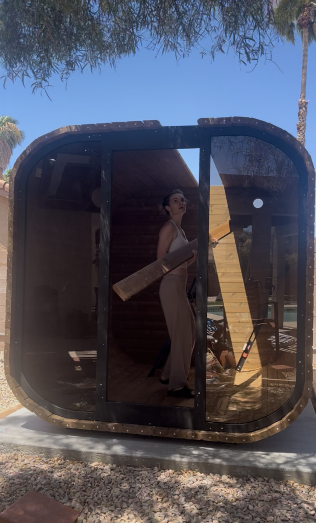 Emily holding a long wooden board inside the partially assembled sauna, viewed through the front opening.
