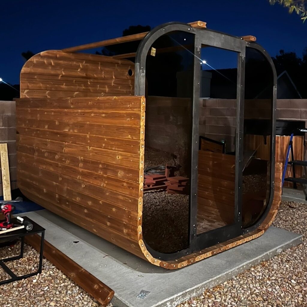 Partially completed wooden barrel sauna in the backyard at dusk, with the walls assembled and roof unfinished.