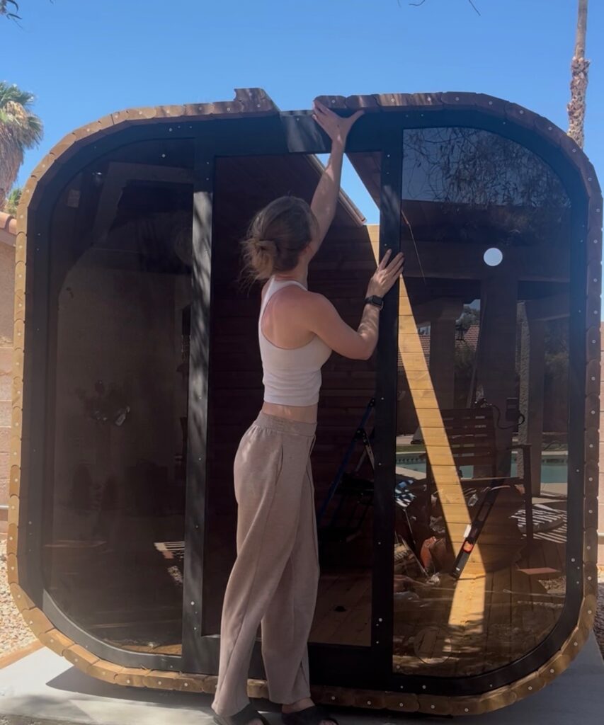 Emily working on the upper section of the wooden sauna, reaching up to position the roof components.
