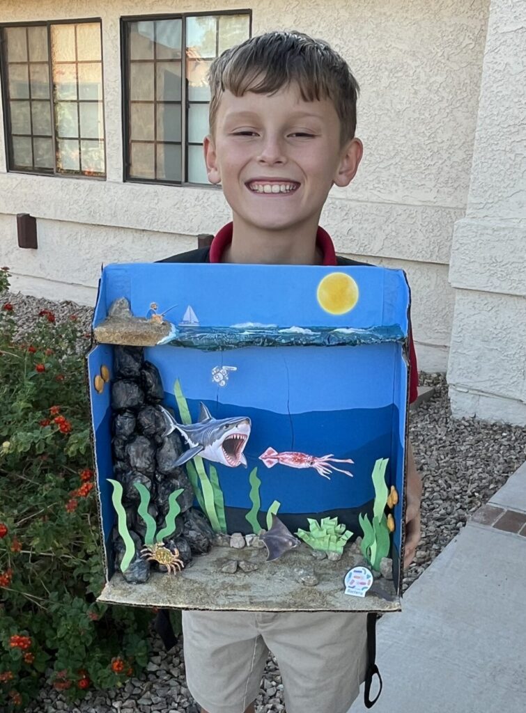 Harrison smiling while holding his handmade ocean habitat diorama featuring a great white shark, squid, rocks, and underwater plants.