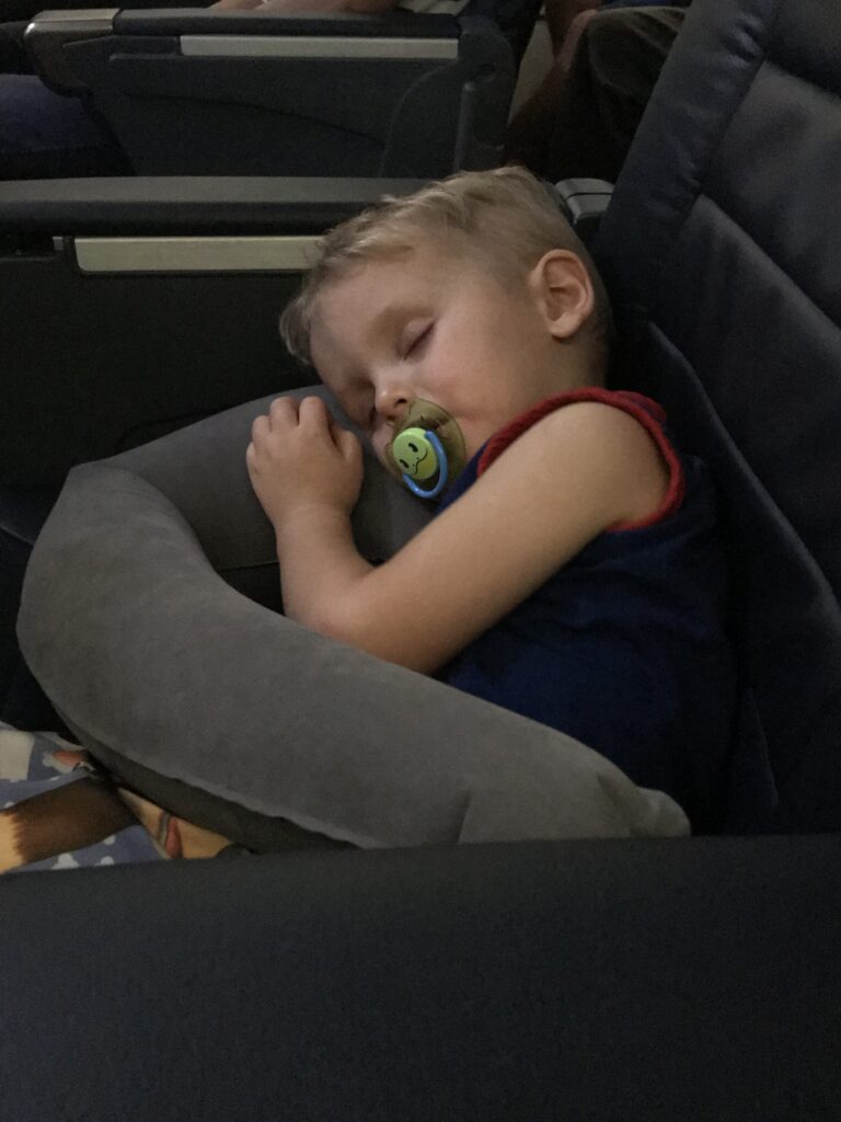 A child sleeps with a pacifier against a gray neck pillow in an airplane seat.