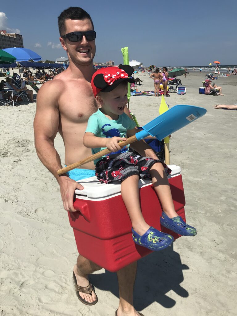 A man carries a red cooler with a child seated on top at the beach.