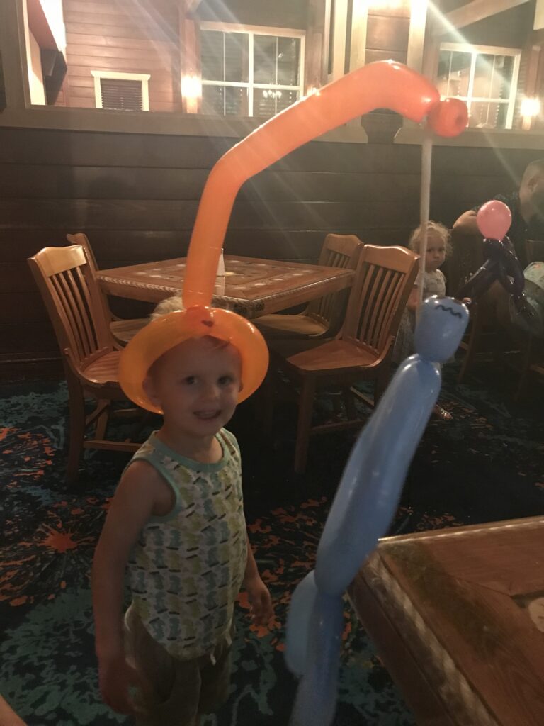 A child wears a tall orange balloon hat and holds a blue balloon creation.