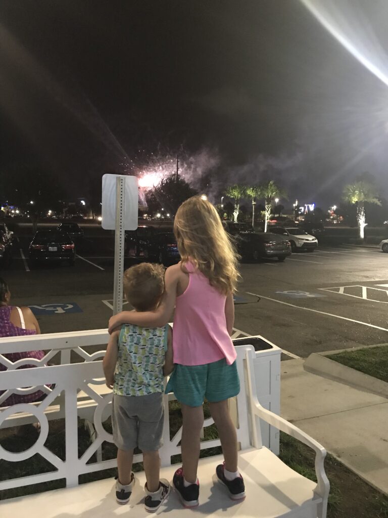 Two children stand at a white railing with fireworks in the night sky.