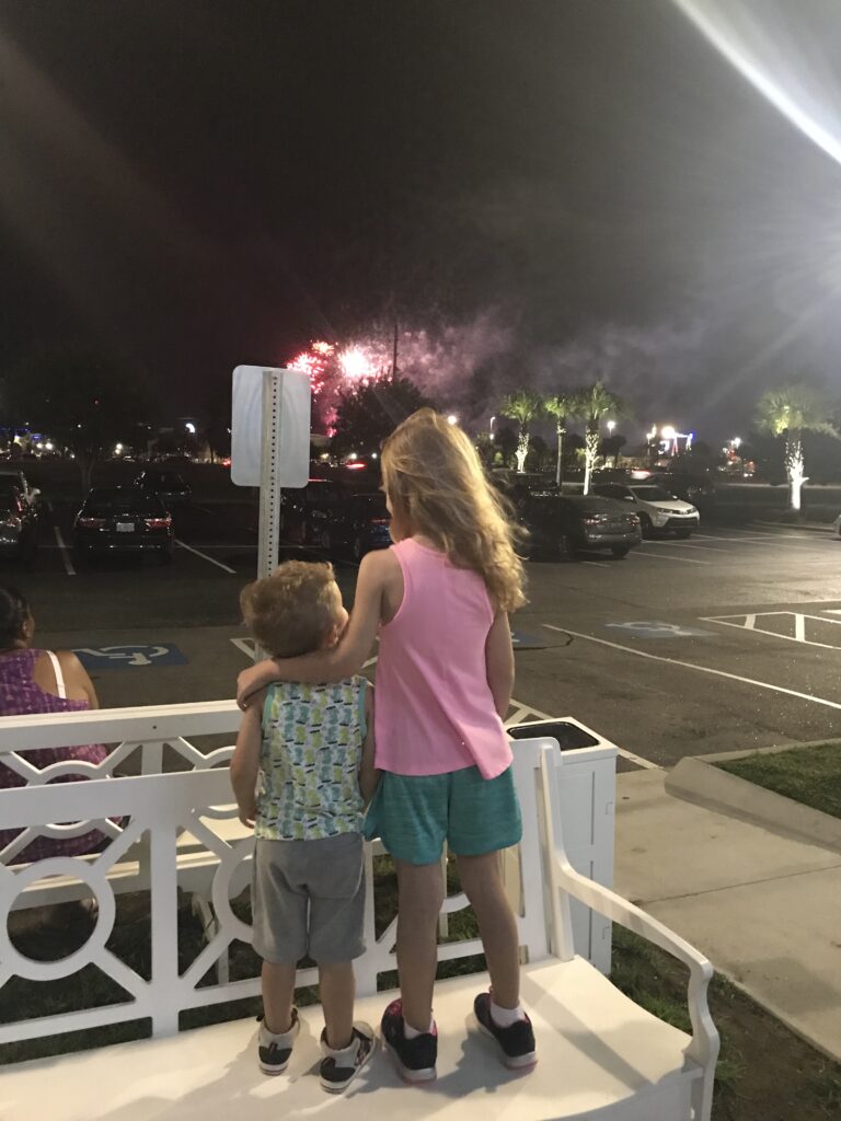 Two children stand together by a white railing watching fireworks at night.