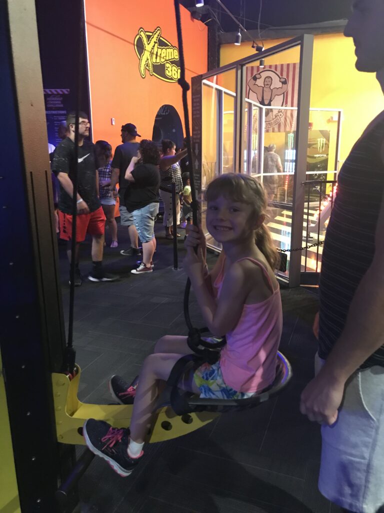 A smiling child holding a rope in a yellow suspended chair, with an adult standing alongside.