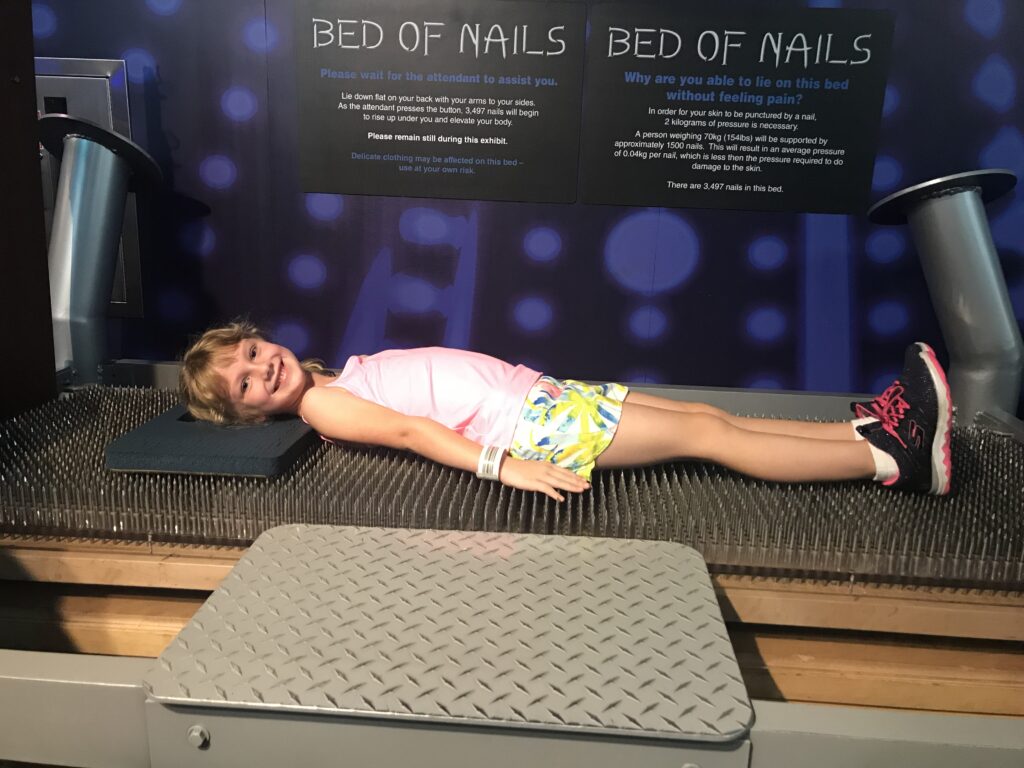 A smiling child lying on a bed-of-nails exhibit beneath explanatory signs.