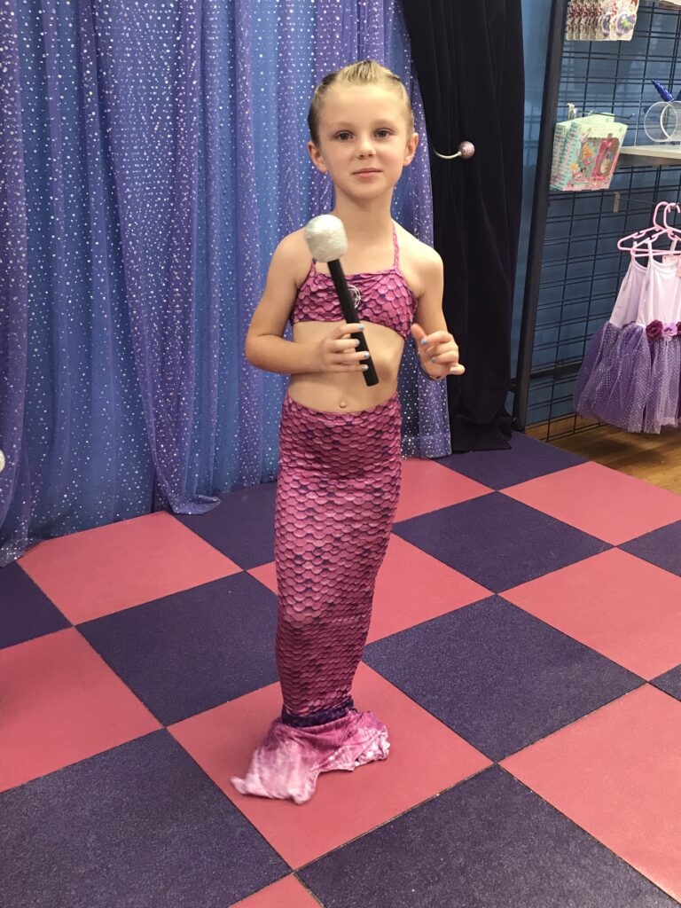 A child in a pink mermaid costume holding a silver-topped microphone in front of a sparkling curtain.