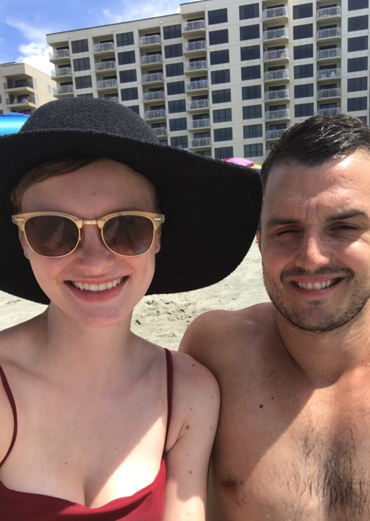 Two adults smile for a beach selfie with buildings behind them.