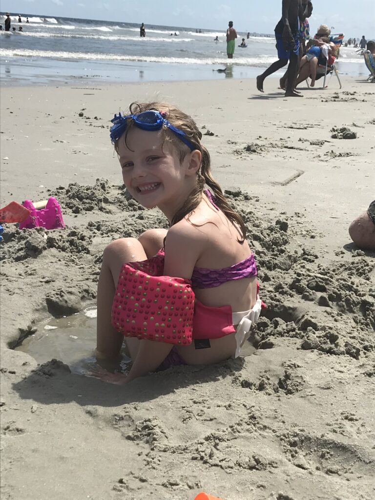 A child wearing pink flotation aids sits in the sand at the beach.