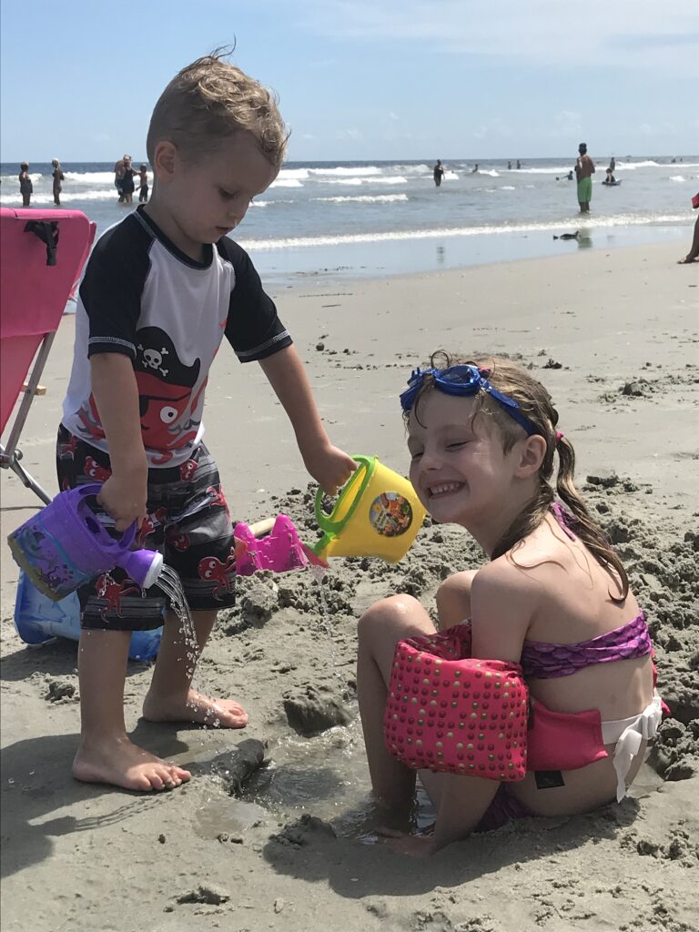 Two children play with colorful buckets in the sand beside the ocean.