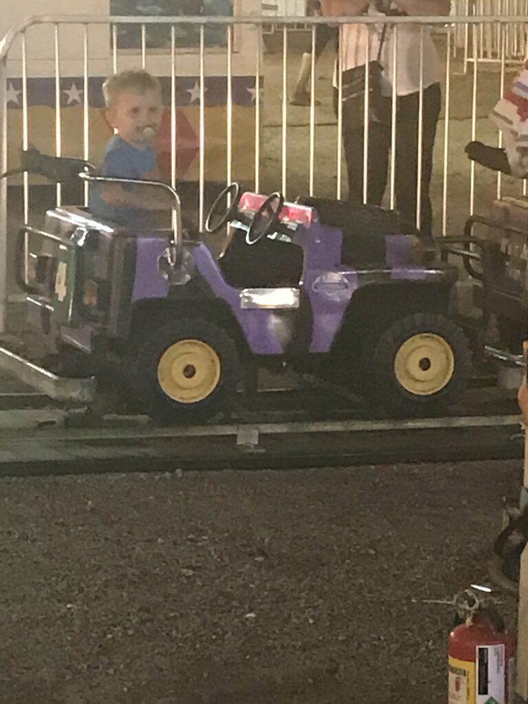A child rides in a purple miniature jeep beside a white fence.