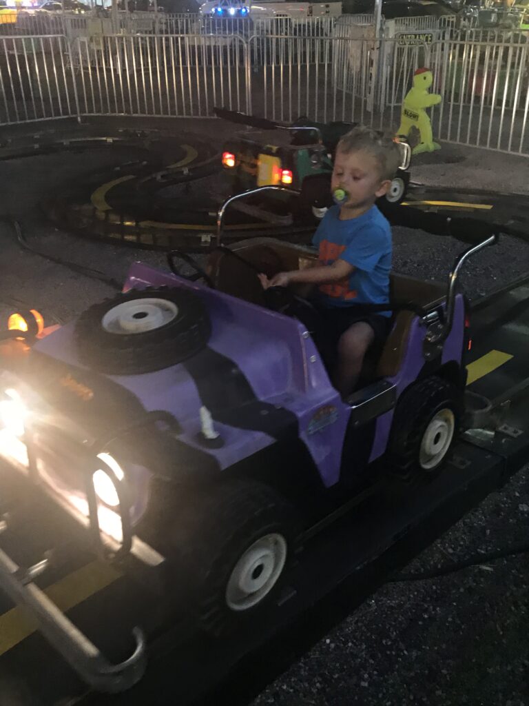 A child sits at the wheel of a purple miniature jeep ride at night.