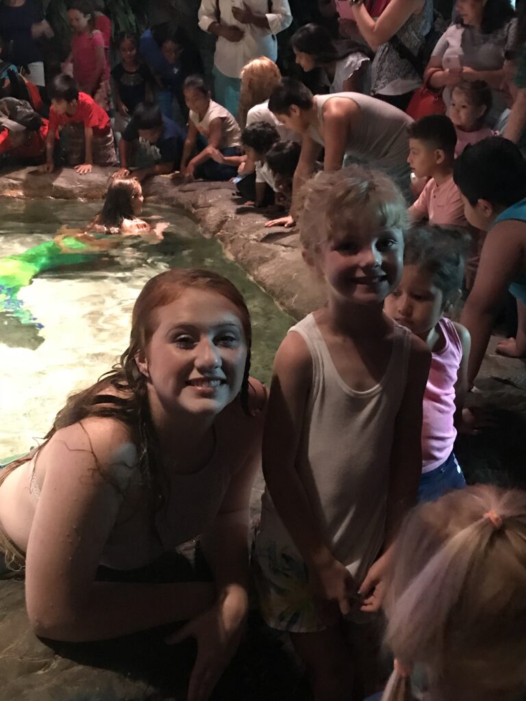 A child poses beside a mermaid performer at the edge of an indoor pool.